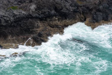 Dalgalar kayalık kıyı şeridine çarpar, köpük oluşturur ve turkuaz suya su sıçratır..
