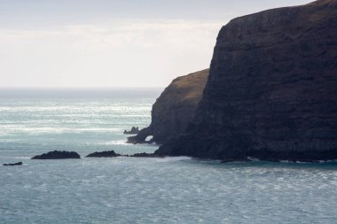 Uçurumları ve kayalık oluşumları olan engebeli bir sahil manzarası. Okyanus dalgaları, güneş ışığını yansıtarak kıyıya çarpar. Gökyüzü biraz bulutlu, sakin bir atmosfer yaratıyor..