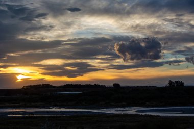 Gün batımında sakin bir manzara, ön planda kıvrılan bir nehir, siluetli ağaçlar, ve bulutlarla dolu dramatik bir gökyüzü ve sıcak turuncu ve sarı renkler..