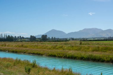 Bereketli yeşil çayırların arasından akan turkuaz bir nehrin, berrak mavi gökyüzünün altındaki uzak dağların bulunduğu sakin bir manzara..