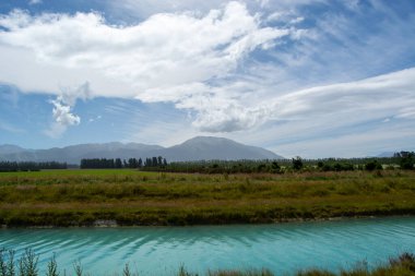 Bereketli yeşil çimenler ve tarlalarla çevrili turkuaz suyu olan sakin bir nehrin olduğu sakin bir manzara. Arka planda, görkemli dağlar parçalı bulutlu bir gökyüzünün altında yükselir ve kırsal bir manzara yaratır..