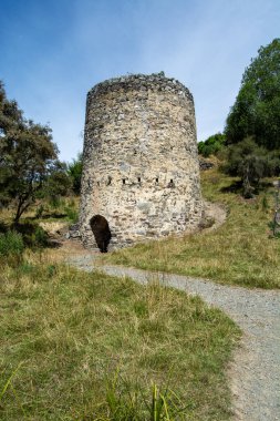 Çimlerle ve yeşilliklerle çevrili tarihi bir taş kule ve ona doğru uzanan toprak bir patika. Kule silindirik ve yıpranmış, açık mavi gökyüzüne karşı kurulmuş..