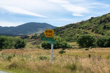 Tarihi bir yere ve fırına giden yolu gösteren sarı bir işaret direğinin olduğu kırsal bir arazi. Arkaplanda yuvarlanan tepeler ve mavi gökyüzünün altında ince bulutlu yemyeşil yeşillikler var..