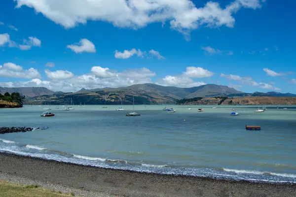 Tepelerle çevrili birkaç teknenin bulunduğu sakin bir körfezin manzarası ve tüylü beyaz bulutlarla dolu açık mavi bir gökyüzünün altında. Kıyı şeridinde çakıl taşları ve yumuşak dalgalar var..