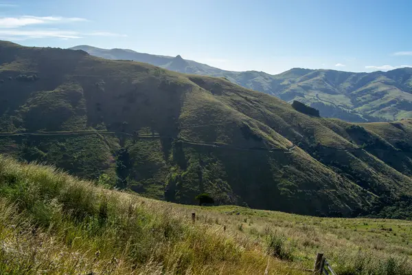 Berrak mavi gökyüzünün altındaki yuvarlanan tepelerin ve dağların manzarası. Çevrede yemyeşil çimenler ve dolambaçlı bir yol bulunur. Doğanın güzelliğini gözler önüne serer..