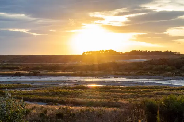 Gün batımında dingin bir manzara, yemyeşil ve çayırlarda kıvrılan bir nehri gösteriyor. Güneş uzak tepelerin ardında batıyor, sahnenin üzerine altın ışınlar saçıyor..
