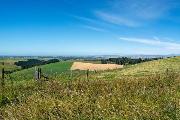 A scenic view of rolling green hills under a clear blue sky, with a patchwork of fields and distant mountains. The foreground features tall grass and a wooden fence.