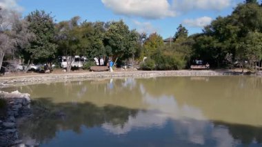 Bereketli yeşilliklerle ve parçalı bulutlu bir gökyüzünün altındaki ağaçlarla çevrili sakin bir gölet. Su, ağaçları ve bulutları yansıtır ve barışçıl bir atmosfer yaratır..