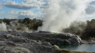 Sıcak kaynaklar ve gayzerlerden yükselen buharlı bir jeotermal arazi, parçalı bulutlu bir gökyüzü altında kayalık arazi ve yemyeşil alanla çevrili..
