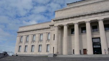 Büyük sütunları ve merdivenleri olan klasik mimarisi olan büyük bir bina. Ön cephe yazıtlarla süslenmiş ve sol tarafında modern bir uzantısı var. Gökyüzü parçalı bulutlu.