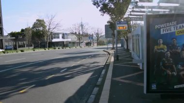 Bir otobüs durağı, açık mavi gökyüzü ve seyrek trafiğin yer aldığı sakin bir şehir manzarası. Yol ağaçlar ve modern binalarla kaplı, sakin bir atmosfer yaratıyor..