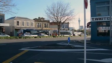 Modern ve tarihi binaların karışımından oluşan bir sokak manzarası. Ön planda yeşil çalılarla kaplı bir yol boyunca park edilmiş arabalar görülüyor. Soldaki modern bina parlak bir tasarıma sahip, sağdaki bina ise klasik mimari sergiliyor..