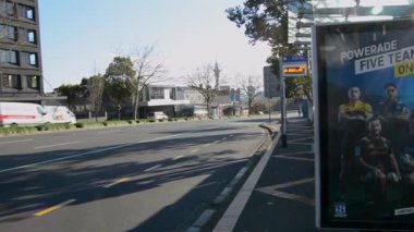 Ağaçlar ve modern binalarla çevrili geniş şeritli, sakin bir sokak manzarası. Gökyüzü açık ve mavi, güneşli bir günü işaret ediyor..