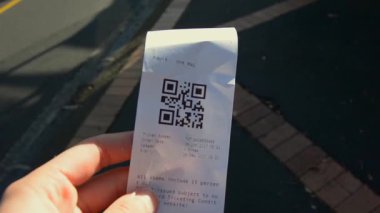 Elinde QR kodlu bir toplu taşıma bileti, basılı detaylar ve kısmen görünür bir sokak geçmişi olan bir el..
