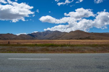 Uçsuz bucaksız manzarada, parlak mavi bir gökyüzü altında tüylü beyaz bulutlarla dağları barındıran bir manzara. Ön planda bir çim tarlası ve bir yol var..