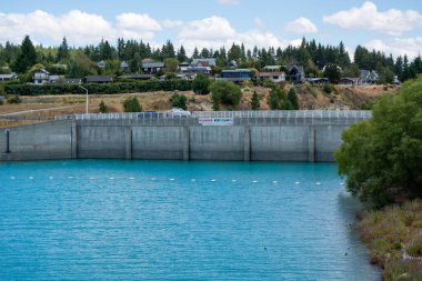 Yeşil ağaçlarla çevrili turkuaz suyu olan bir baraj manzarası ve parçalı bulutlu bir gökyüzünün altındaki dağ yamacında evler..