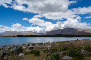 Etrafı kayalık kıyılar ve yemyeşil yeşilliklerle çevrili sakin mavi bir göl manzarası. Arka planda, parlak mavi bir gökyüzünün altında kabarık beyaz bulutlarla süslenmiş görkemli dağlar yükselir..