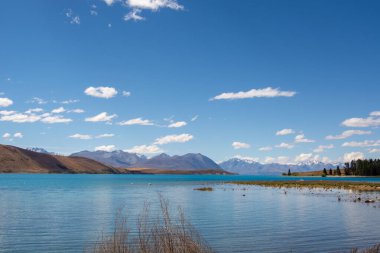 Pofuduk beyaz bulutlu, parlak mavi gökyüzünün altında dağlarla çevrili berrak mavi bir göle sahip huzurlu bir manzara. Ön planda uzun otlar var ve uzak kıyı ağaçlarla kaplı..