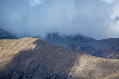 Kısmen bulutlarla kaplı engebeli dağların manzaralı manzarası, dünyevi tonların ve dramatik ışıklandırmanın bir karışımını sergiliyor..