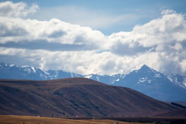Parçalı bulutlu bir gökyüzünün altında kar kaplı tepeleri olan dağların manzarası. Ön planda gökyüzü ve dağların mavi ve beyaz ile çelişen kahverengimsi bir tonu ile yuvarlanan tepeler yer almaktadır..