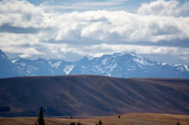 Bulutlu bir gökyüzünün altında karla kaplı tepeleri olan dağların manzarası. Ön planda engebeli tepeler ve seyrek bitki örtüsü yer alıyor ve arka plandaki engebeli sıradağlarla bir tezat oluşturuyor..
