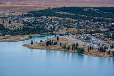 Ağaçlar ve açık arazilerle çevrili, göl kenarındaki küçük bir kasabanın havadan görünüşü. Su manzarayı yansıtır, sakin bir atmosfer yaratır..