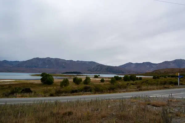 Bulutlu bir gökyüzünün altında dağlarla çevrili bir göl manzarası. Ön planda seyrek bitki örtüsü ve gölün kıyısında uzanan ağaçlarla dolu bir yol yer alıyor..