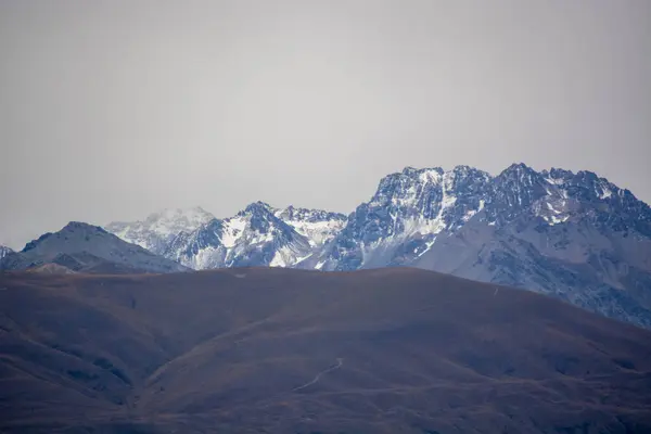 Bulutlu bir gökyüzünün altında karla kaplı tepeleri olan engebeli dağların panoramik görüntüsü. Ön planda, sessiz renk paleti ile yuvarlanan tepeler, sakin ve dramatik bir manzara yaratıyor..