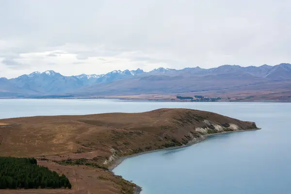 Sakin bir göle sahip sakin bir manzara hafif eğimli bir tepe ve bulutlu bir gökyüzünün altındaki uzak dağlarla çevrili..