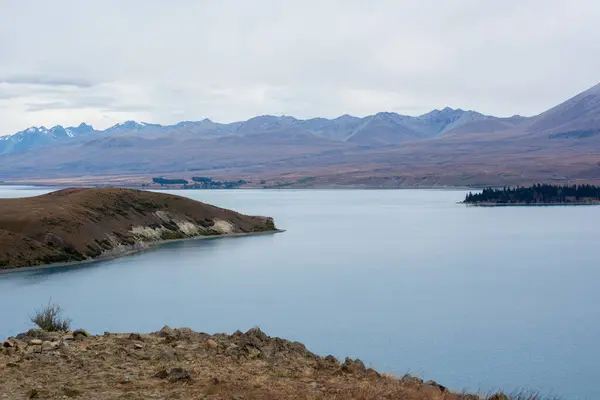 Dağlar ve yuvarlanan tepelerle çevrili sakin bir göl içeren sakin bir manzara. Gökyüzü bulutlu, sakin bir atmosfer yaratıyor. Su, manzaranın soluk renklerini yansıtır..