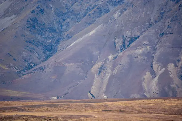 Kuru, kayalık yamaçları ve seyrek bitki örtüsüyle engebeli dağları barındıran engin bir manzara. Arazi toprak tonları ve izolasyon hissi ile karakterize edilir, sakin ama sert bir çevreyi çağrıştırır..