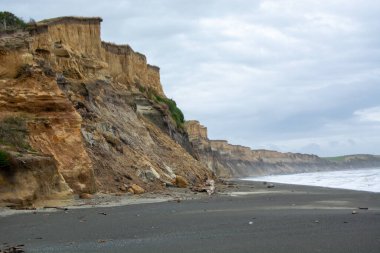 Katmanlı kaya oluşumları, kumlu plaj ve bulutlu bir gökyüzünün altında dalgalı okyanus dalgaları olan sarp kayalıkların yer aldığı engebeli bir sahil şeridi..