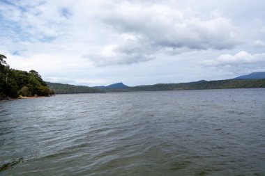 Bereketli yeşil tepeler ve bulutlu bir gökyüzünün altındaki dağlarla çevrili sakin bir göl manzarası. Su sakin, manzarayı yansıtıyor, barışçıl bir atmosfer yaratıyor..