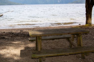 Etrafı doğa ile çevrili sakin bir göl kenarındaki kırsal ahşap piknik masası. Durgun su, arka plandaki dağları yansıtır ve sakin bir açık hava ortamı yaratır..