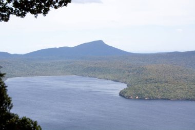 Önünde sakin bir su kütlesi olan dağlık bir manzaranın manzarası. Bölgeyi çevreleyen yemyeşil tepeler ve ağaçlar, parçalı bulutlu bir gökyüzünün altında.
