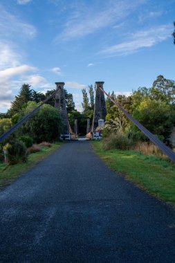 Yeşillik ve açık mavi gökyüzü ile çevrili taş kuleleri olan asma bir köprünün manzarası. Köprüye giden patika çimen ve ağaçlarla kaplı. Huzurlu bir atmosfer yaratıyor..