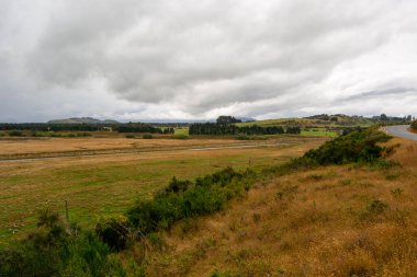 Bulutlu bir gökyüzünün altında çimenli bir tarlanın yer aldığı engin bir manzara. Sahnede uzak tepeler ve kıvrımlı bir yol, yeşil alanlar ve ön planda kuru otlar yer alıyor..