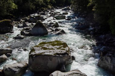 Büyük kaya parçalarının kısmen suya gömüldüğü kayalık bir arazide akan bir nehrin manzarası. Güneş ışığı sudan yansıyarak sakin bir atmosfer yaratır..