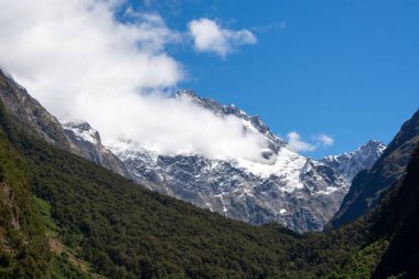 Bereketli yeşil vadilerle çevrili karla kaplı dağların çarpıcı manzarası. Parlak mavi gökyüzü altında dağınık bulutlar..