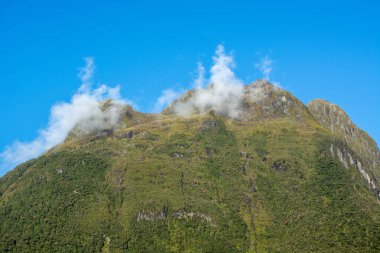 Bereketli yeşilliklerle kaplı görkemli bir dağ zirvesi. Zirvenin etrafında süzülen ince bulutlar berrak mavi gökyüzüne karşı..