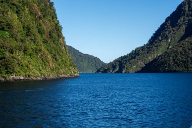 Berrak mavi bir gökyüzünün altında yemyeşil dağlarla çevrili derin mavi bir göl manzarası. Manzara sarp kayalıklara ve canlı bitki örtüsüne sahip. Huzurlu bir doğal ortam yaratıyor..