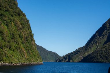 Sabit yeşil tepelerin bulunduğu sakin bir manzara berrak mavi bir gökyüzünün altında sakin mavi bir su geçidini kuşatıyor. Sahne, rahatlama ve macera temaları için ideal olan doğanın sükunetini yakalıyor..