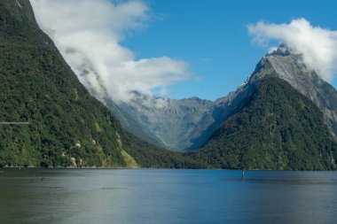 Büyüleyici bir fiyort manzarası yüksek dağlar ve yemyeşil ile çevrili, bulutlar kısmen zirveleri kaplıyor ve ön planda sakin bir su kütlesi var..