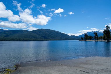 Berrak mavi sularla çevrili dingin bir göl manzarası, yemyeşil dağlar ve kabarık beyaz bulutlu parlak mavi bir gökyüzü. Ön planda beton bir kıyı yer alıyor.