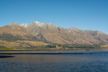 Etrafındaki dağları yansıtan sakin bir göl içeren sakin bir manzara. Dağlar kısmen karla kaplı, yukarıda açık mavi bir gökyüzü var. Ön planda sakin bir su yüzeyi görünüyor. Barışçıl atmosferi güçlendiriyor..