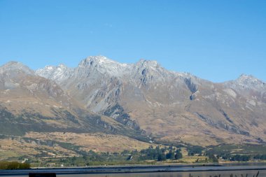Berrak bir mavi gökyüzünün altındaki kar kaplı dağların çarpıcı manzarası, kayan tepelerin ön planı ve manzarayı yansıtan sakin bir göl..