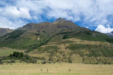 Yeşil tepeleri ve bulutlarla benekli mavi gökyüzü ile dağlık bir manzaranın manzarası. Ön planda çimenlik bir alan yer alırken, arka planda belirgin bir dağ zirvesi yer alıyor.