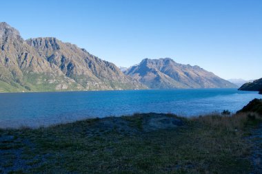 Berrak mavi gökyüzünün altında görkemli dağlarla çevrili sakin bir göl manzarası. Su dağları yansıtır, resim gibi bir manzara yaratır..