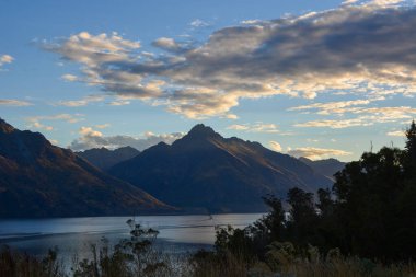 Gün batımında bulutlu bir gökyüzünün altında dağları ve sakin bir gölü barındıran sakin bir manzara. Sahne, doğanın güzelliğini, yumuşak ışık ve suda yansımalarla yakalar..