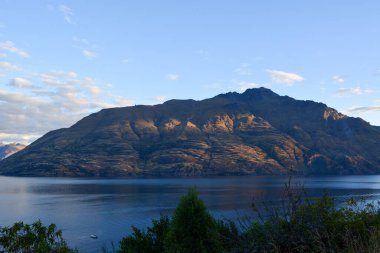 Berrak mavi gökyüzünün altında sakin bir göle yansıyan sakin bir dağ manzarası. Güneş ışığı, doğal güzelliğini artırmak için dağa yumuşak gölgeler bırakır..